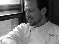 A black and white close-up of former Season 6 Next Food Network Star Brook Harlan wearing a white chefs jacket while seated and looking out a window
