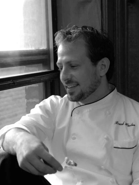 A black and white close-up of former Season 6 Next Food Network Star Brook Harlan wearing a white chefs jacket while seated and looking out a window