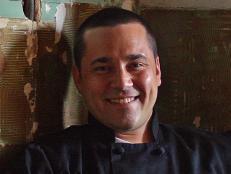A close-up of former Season 6 Next Food Network Star Teddy Folkman weaing a black chefs jacket while seated and holding a beverage