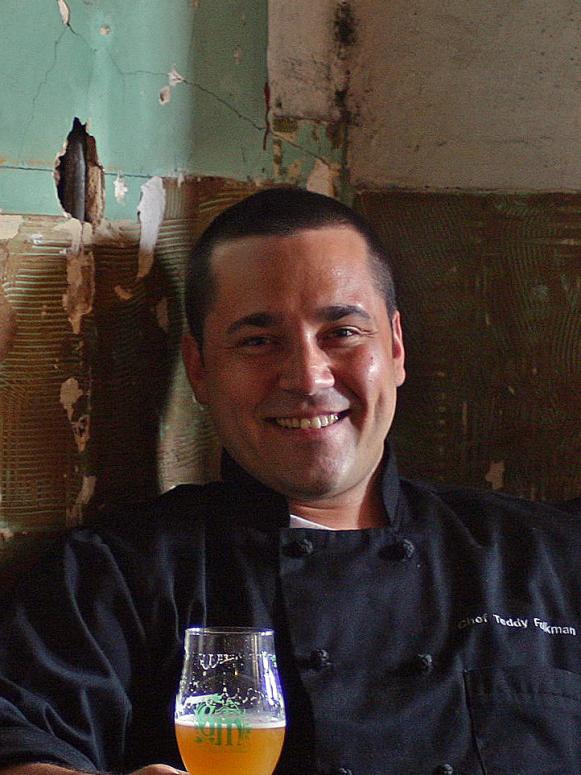 A close-up of former Season 6 Next Food Network Star Teddy Folkman weaing a black chefs jacket while seated and holding a beverage