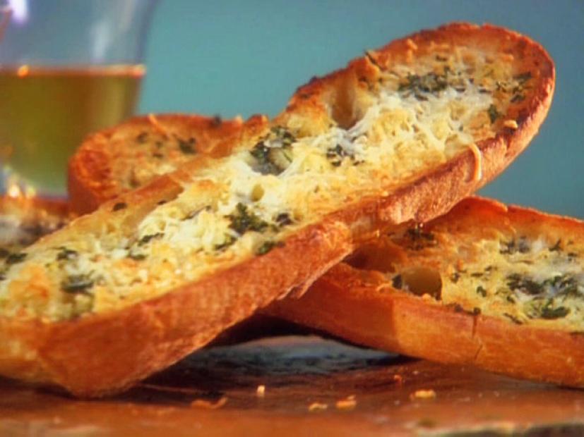 Halved baguettes topped with butter, garlic, parsley and asiago cheese which are crisscrossed on a stone like surface