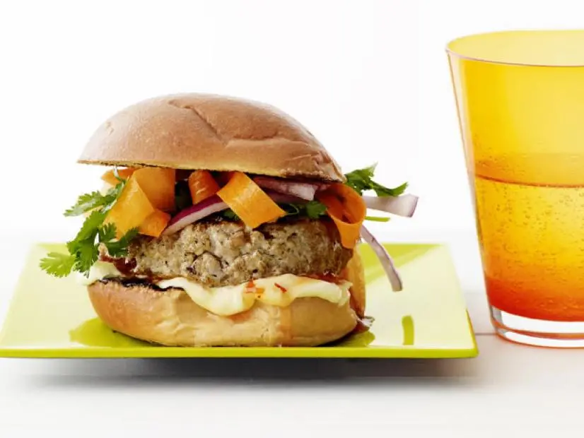 A chicken sandwich topped with herbs, carrots and red onions on a small square light green plate