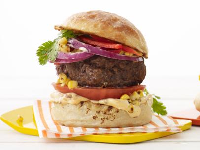 A hamburger topped with herbs, corn, peppers and red onions on a square yellow plate
