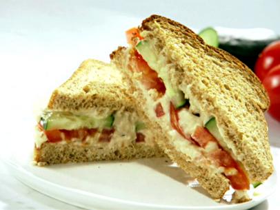 A hummus sandwich made with white beans and topped with slices of cucumber and tomatoes. This is served on toasted wheat bread that has been cut in half diagonally and placed on a white plate.