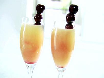 Two champagne glasses that have been filled with ingredients to make a venice sunset cocktail. These were made with a cherry sauce, sparkling white grape juice, and grapefruit juice. Cherries were placed on skewers for garnish.