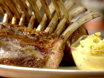 A close up of lamb chops that have been cooked and are standing up against each other. The bones of the lamb chops are in between each other to help them to stand up. A small clear glass bowl has the pineapple mint in it. The bowl and lamp chops arer sitting on a white plate.
