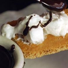 Close up of chocolate sauce being poured over marshmallow on a graham cracker, making S'mores. 