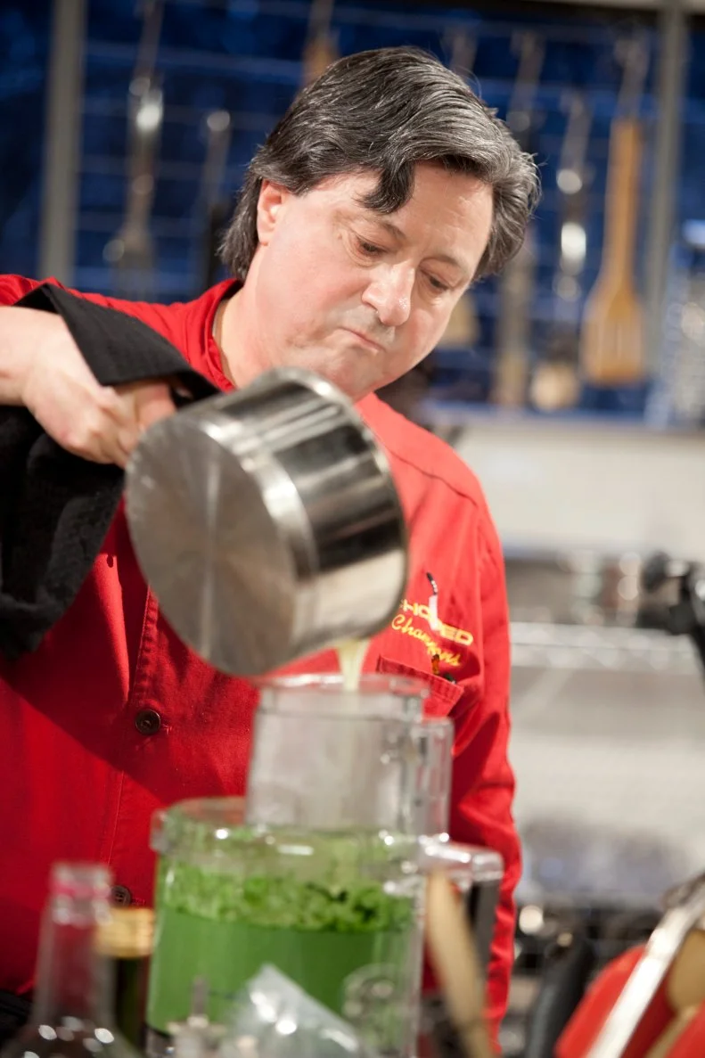 Chef Jean-Louis Gerin pours liquid into a food processor to make a sauce, as seen on Food Network’s Chopped Champions, Season 14.
