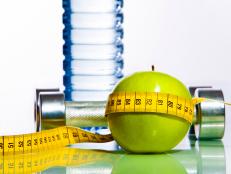 Green apple bounded with measuring tape with two dumbbells and bottle of water behind