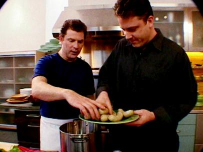 Bobby Flay and a friend place bratwurst into a pot with other ingredients to simmer.
