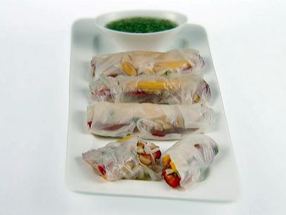 A white rectangular platter with fruit spring rolls. The one in front has been cut in half to show what it looks like on the inside. They were filled with pad thai noodles and mint, strawberries, mango, toasted almonds, and wrapped with rice paper. There is a dipping sauce in a bowl.