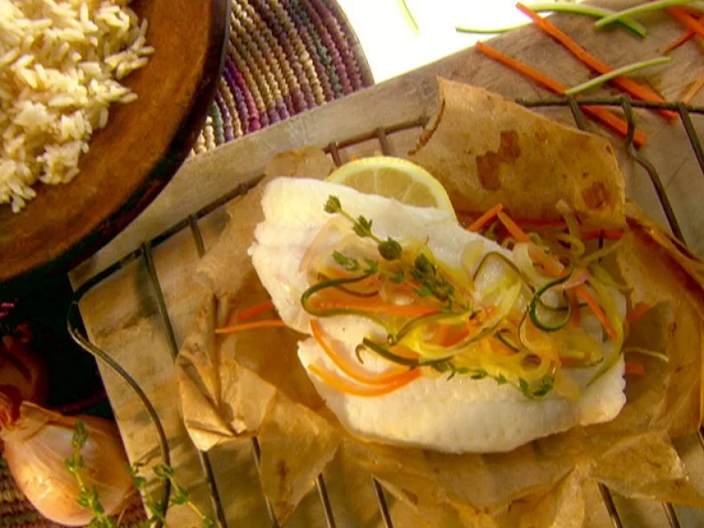 A view of fish en papillote on a brown piece of paper. The fish has lemon, julienned carrots, green zucchini and red onions. The paper is sitting on a light tan wood surface. There is a dark brown and black bowl of brown rice sitting next to the fish. There are some of the julienned carrots and zucchini sitting on the wood surface beside the fish. 