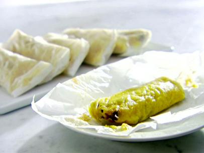 A white plate with a bean and cheese tamale. There are more wrapped bean and cheese tamales on a platter in the background.