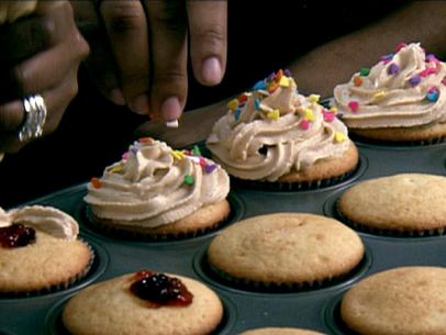 A pan of peanut butter and jelly cupcakes. Some of them have been filled with jelly and topped with the peanut butter cream cheese frosting and sprinkles while other just have the jelly and some do not. 