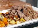 Cooked beef chuck roast sits on a white plate with other vegetables. The vegetables are carrots, onions and potatoes that have been roasted in an oven. The roast has been shredded into small pieces.