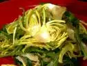 A close up of a salad on a red plate. This salad was made with shaved artichoke, arugula, and parmigiano cheese. It has been dressed with finishing oil.