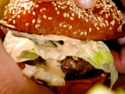 A close up of hands as they hold a prime beef burger with lettuce, tomato, pickles; pickle mayo and melted cheese.