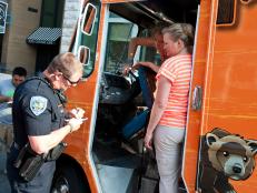 A Fayetteville police man talks with Momma's Grizzly Grub's Angela Reynolds and Tiffany Seth following an accident with her truck and a lamp post in Fayetteville, Ark., as seen on Food Network's The Great Food Tuck Race, Season 3.