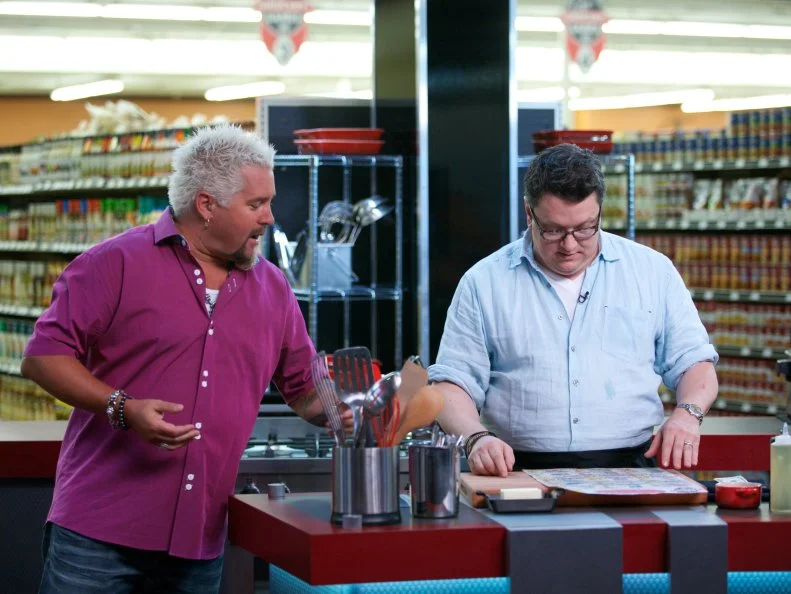 Host Guy Fieri with chef Tom Ramsey during the cooking part of Game 2, Budget Battle, Family Meal for 4 with $5.91, as seen on Food Network's Guy's Grocery Games, Season 1.