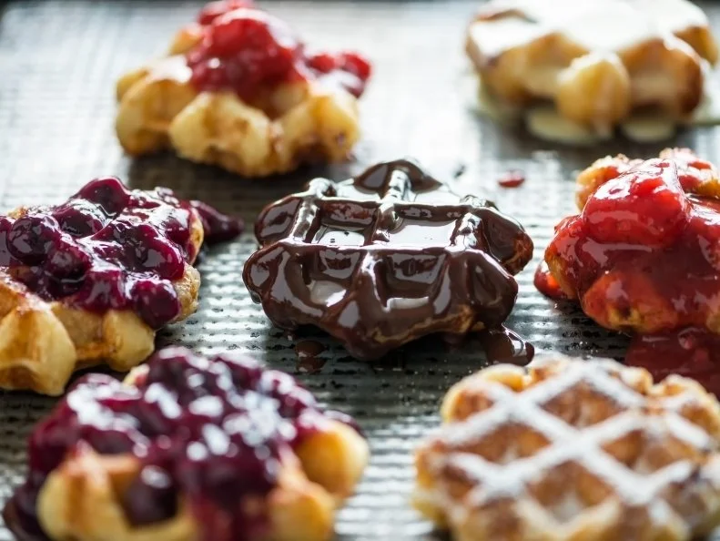 Old baking pan full of bite sized homemade Belgium Liege waffles randomly arranged, and decorated with blueberry and strawberry sauce (compote), dark chocolate ganache, white chocolate ganache, and icing sugar.  Drips, spills and a little messy.  Closeup view