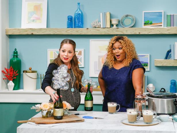 Host Marcela Valladolid demonstrates an idea for crafting a centerpiece with a leftover champagne bottle for fellow hosts Sunny Anderson and Katie Lee during a segment on Lazy Brunch, as seen on Food Network's The Kitchen, Season 11.