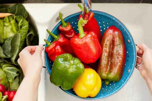 Directly above cropped image of hands washing bell peppers in colander. There are various vegetables on sink. Woman is holding fresh vegetables in container under faucet. She is in domestic kitchen.