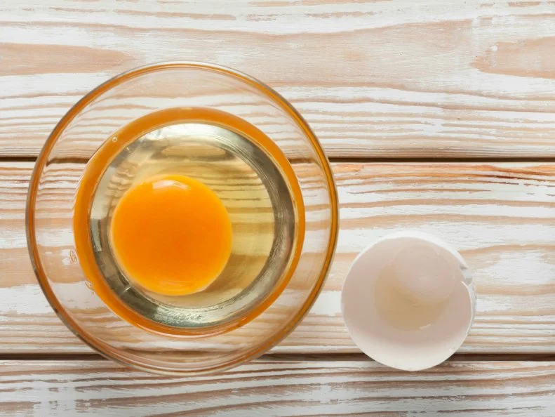 Top view of yolk and egg white in glass bowl on white weathered wooden table
