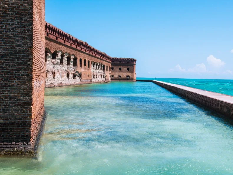 This is a horizontal, color toned photograph of the historic brick fortress, Fort Jefferson on Garden Key in the Dry Tortugas National Park in Florida. A wall surrounds the former military outpost. Tropical colored salt water fills the moat and surrounding wall.