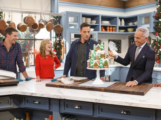 Host Geoffrey Zakarian adds icing to the Gingerbread House Cookie Cake as fellow hosts Jeff Mauro and Sunny Anderson and guests Josh Snyder and Angela Kinsey look on as seen on The Kitchen, Season 15.