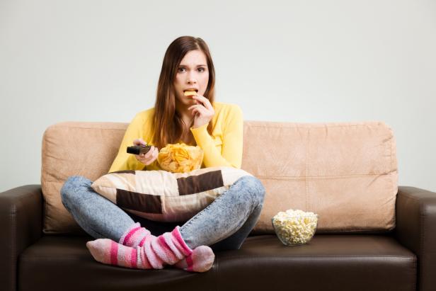 Young woman spends his free time watching TV on the couch at home, munching chips and popcorn. Gray background, easy to remove.