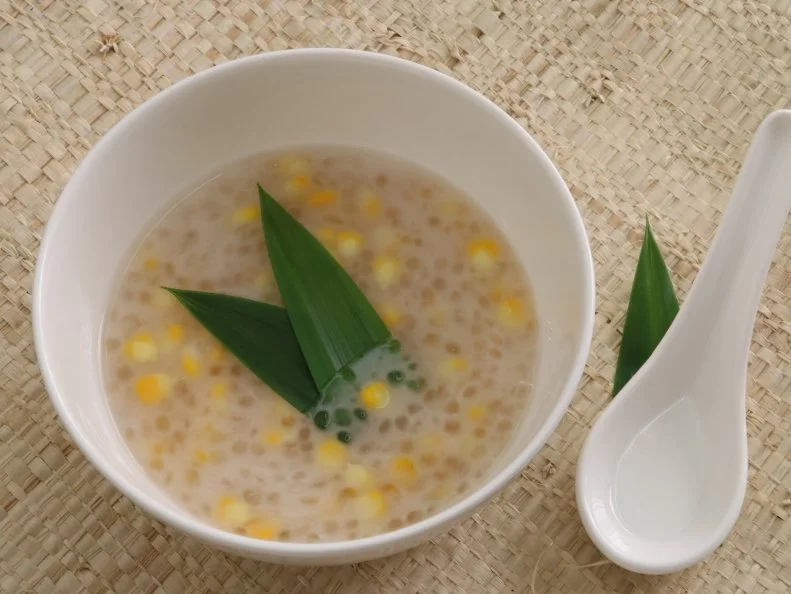 Che Bab, a Vietnamese dish of sweet soup made of tapioca pearls mixed with corn kernels and coconut milk, infused with aromatic pandanus leaf. The paleness of the translucent tapioca pearls is balanced with the bold yellow of the corn. The coconut milk give a creamy flavor and also acts as contrast to the translucent pearls. A white ceramic bowl with minimalist design is used to hold the soup. Pieces of pandanus leaves are used to garnish the soup and also underline the asian style ceramic soup spoon. A woven mat made of organic material is used as the place mat. The image was taken indoor near the window to benefit the sunlight as the natural lighting source.
