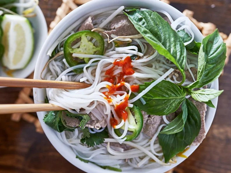 eating beef pho with spicy sriracha sauce shot top down on wooden table with chopsticks
