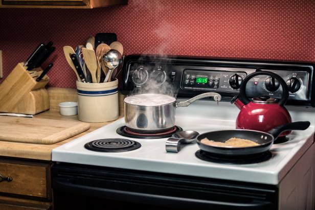 A boiling, steaming, bubbling, overflowing water family size pasta pot and two pieces of fish sizzling in a frying pan are cooking on the stove top burners of a cozy home kitchen electric oven range.