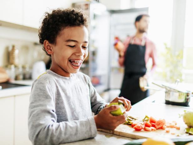 A young boy laughing while he helps his dad prepare lunch by peeling some fruit and veg.
