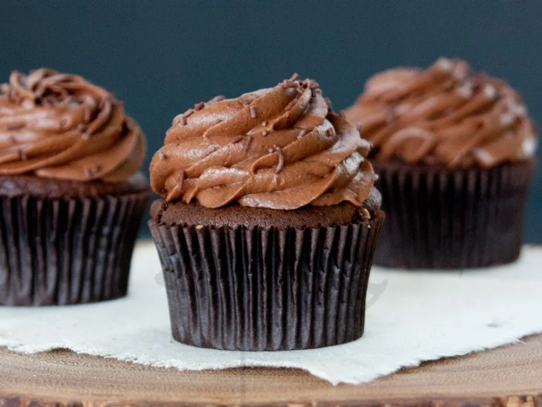 Three chocolate cupcakes with chocolate icing and chocolate sprinkles on cloak on a wood slice witha  black background