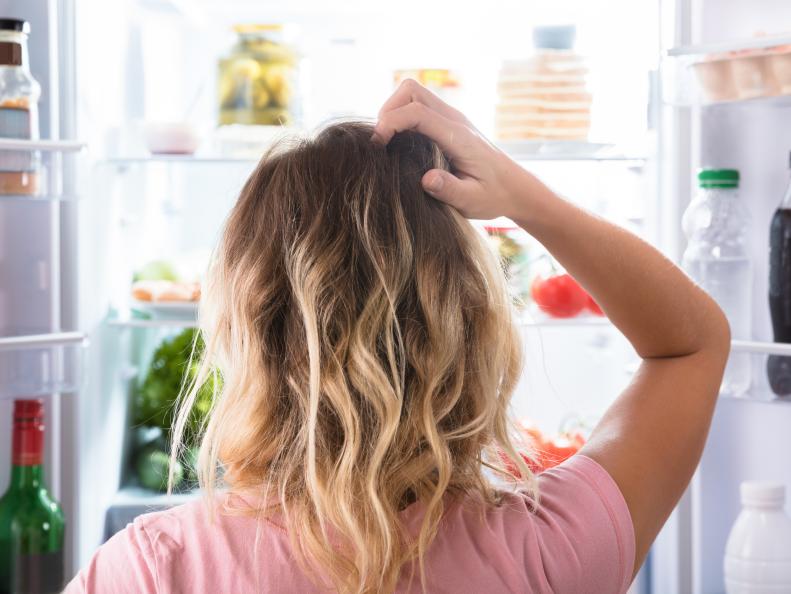Rear View Of A Confused Woman Looking In Open Refrigerator At Home