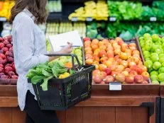 Young woman carries a shopping basket filled with fresh produce. She is shopping for fresh fruit and vegetables in a grocery store.