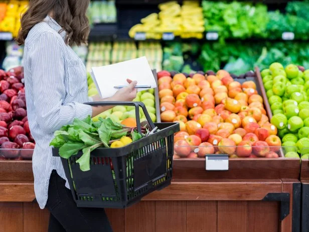 Young woman carries a shopping basket filled with fresh produce. She is shopping for fresh fruit and vegetables in a grocery store.