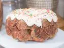 Pull Apart Confetti Cake was prepared by Food and Lifestyle Expert, Brandi Milloy. She cuts store-bought biscuit dough into quarters and places them all inside of a ziplock bag. Chef Stuart O'keeffe sprinkles 3 tablespoons of sprinkles into the bag along with 1/2 cup of granulated sugar, 1/2 cup of brown sugar, and a little bit of cinnamon. He then flipped the bag around multiple times until all the added ingredients were distributed onto the dough pieces. Spray a 10-inch round bundt pan with nonstick cooking spray and pour the covered dough pieces inside. Food and Lifestyle expert Brandi Milloy then begins to make a topping by starting with a little butter in a saucepan and adding sugar. She then mixes it and melts it down until the sugar dissolves. Then add a little almond extract and pour the mixture on top of the dough. Bake for 30 minutes at 350 degrees. After 30 minutes, remove the cake, place foil on top, and bake for an additional 10 minutes. After removing from the oven, flip the cake over onto a pan. Next create a frosting by mixing together 1/2 stick of butter and 1/2 block of cream cheese (about 4 oz.). Add 1 cup powdered sugar, a pinch of salt, 2-3 tablespoons of milk, a dash of vanilla, and mix them all together. Place the frosting on top of the cake, allowing it to fall down the sides. Finish by topping the icing off with sprinkles, as seen on Let's Eat, Season 1.