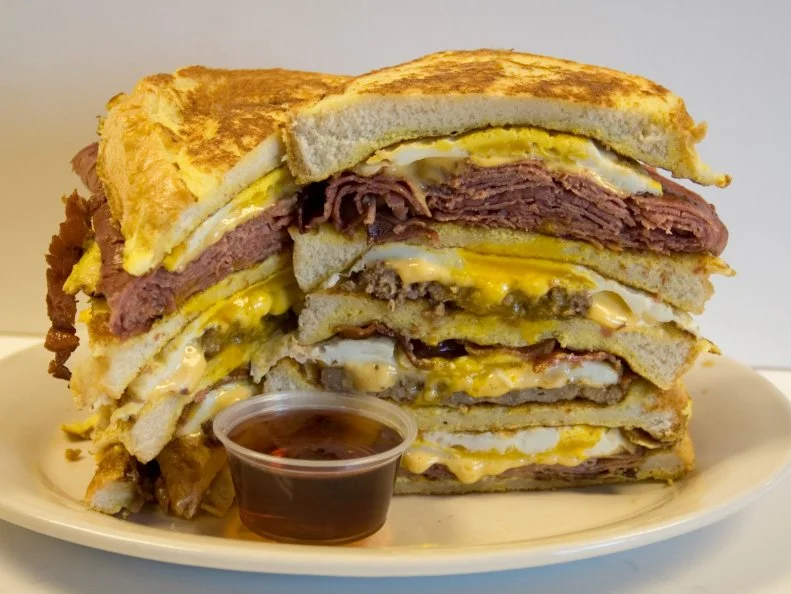 This third-generation Burlington icon is famed for its Texas dogs topped with fiery beef chili, but the breakfast sandwiches are something of a local legend as well. Residents of the Green Mountain State have been known to wax poetic when naming the virtues of the $4.50 McHandy, a no-fuss egg and cheese on a twice-toasted English muffin with a choice of bacon, ham or sausage. Inspired by the formidable appetite of a former UVM student and regular customer, The Super G is a real triple threat. Composed of ham, bacon and sausage, this hefty sandwich piles all that meat plus two eggs and white American cheese onto a round roll from neighboring bakery Koffee Kup. Owner Earl Handy always stabs a fork through the center before delivering The Super G to any of his customer. “If you don’t,” he explains, “it’s gonna fall apart.”
https://handyslunch.files.wordpress.com/2013/10/menu2017.png