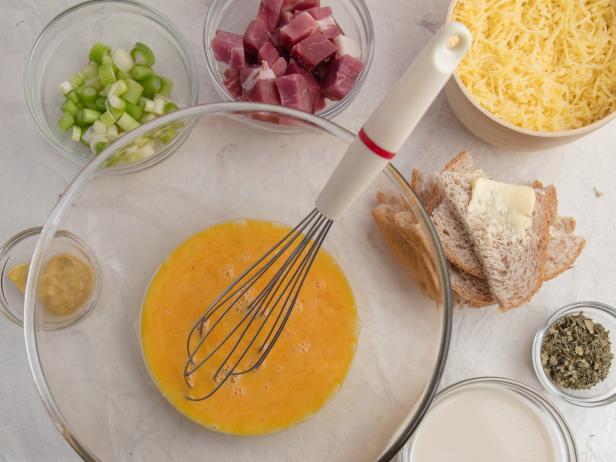 Mise en place for pressure-cooked ham and melted cheese pudding dish. Ingredients are buttered dry brown bread, cup of milk, lightly beaten eggs, tablespoon mustard, dried parsley, grated GruyÃ¨re, sliced scallions and diced cured ham. High point of view.