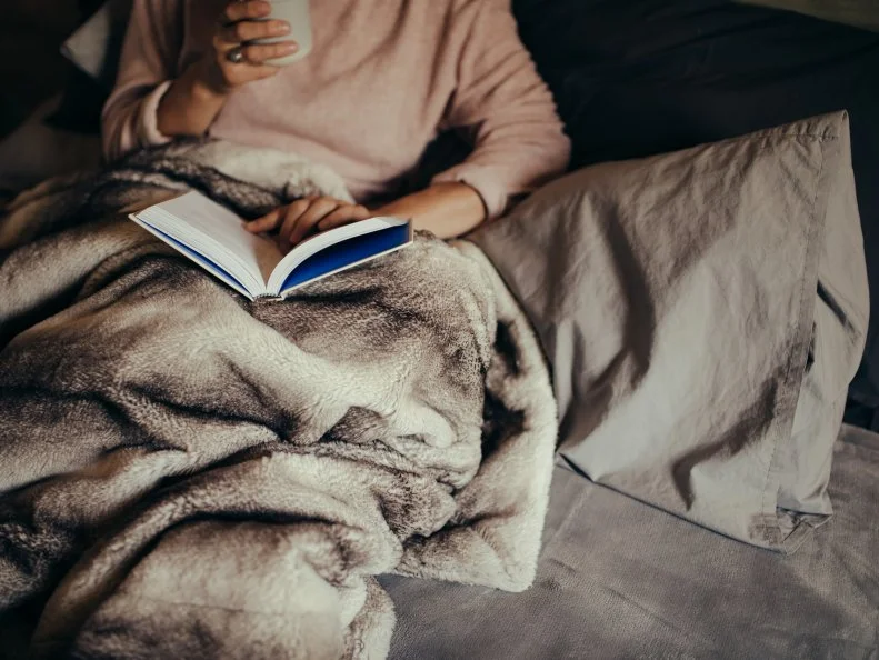 Cropped shot of female on bed reading book and drinking coffee. Female reading book on bed at home.