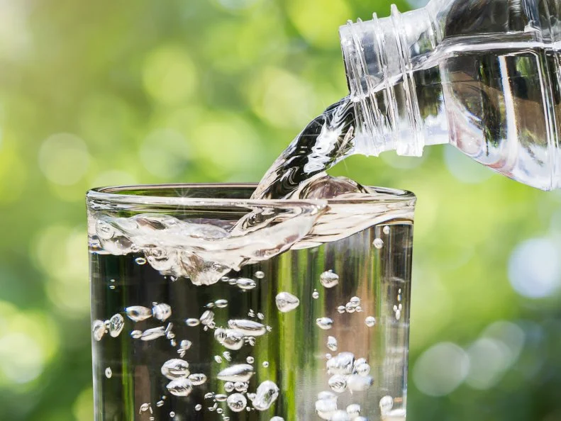 Close up of water flowing from drinking water bottle into glass on blurred green nature bokeh background, healthy drinking clean water concept