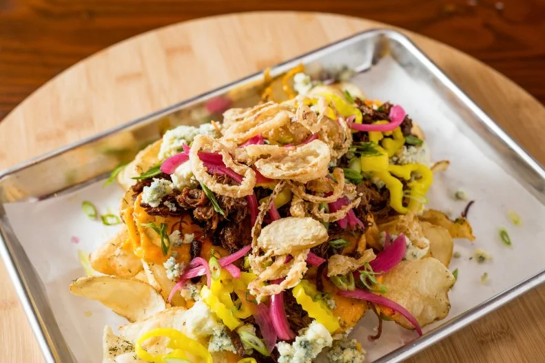 Scott Hall, chef of Bone-In-BBQ in Columbia’s up-and-coming BullStreet District, imbues his recipes with whimsy. There’s the bacon-wrapped mac and cheese, for instance, and the fried chicken and waffle club sandwich. His nachos, Bone Chips, get the same treatment: Hickory-hoisin smoked beef brisket, melted pimento cheese and pickled onion are gussied up with blue-cheese crumble, banana peppers, crispy fried onion and scallions.