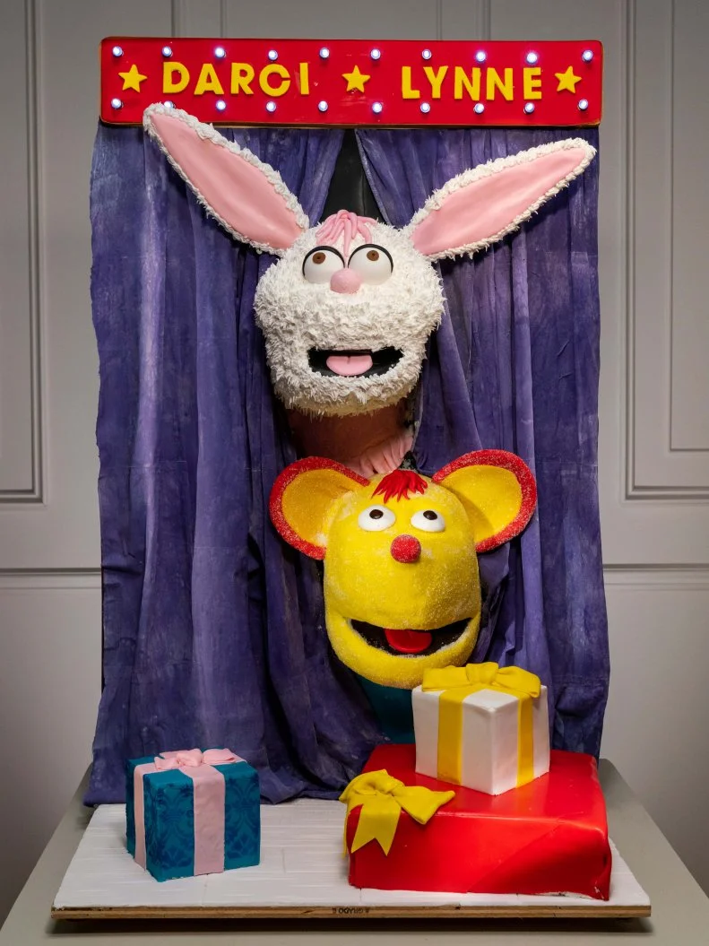 Competitors Matthew Wohlkinger and Sharon Spradley's cake Darci Lynn's puppets, Petunia and Oscar, make their debut on stage in cake, sneaking, from around purple curtains made of treated wafer paper, during the Cake Challenge, as seen on Winner Cake All, Season 1.