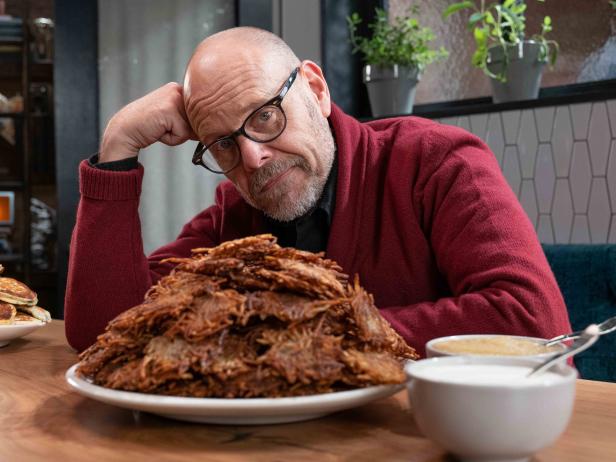 Good Eats: The Return host Alton Brown sits at the kitchen table with a large pile of Potato Latkes for the “Whole Latke Love” episode.
