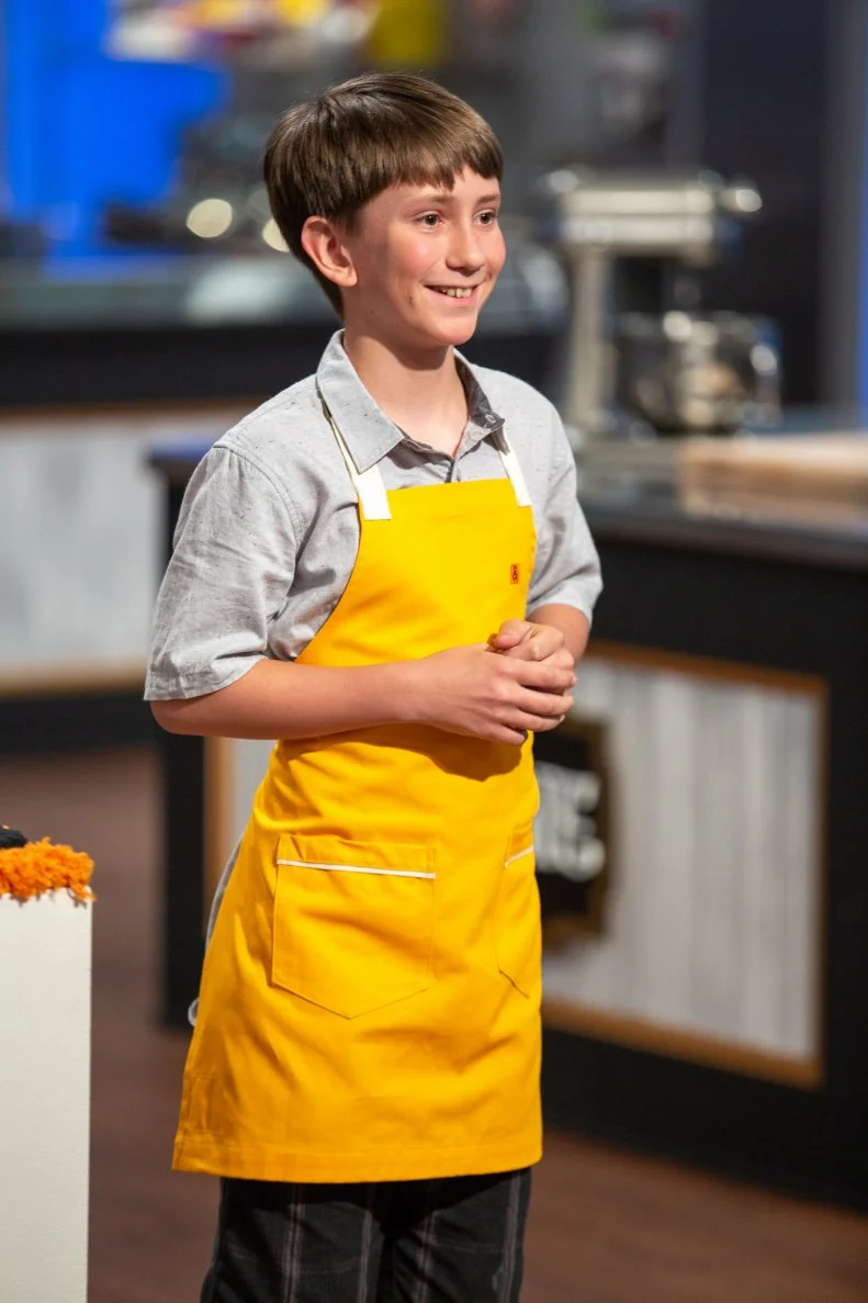 Reggie standing in front of the judges table, as seen on Kids Baking Championship, Season 8.