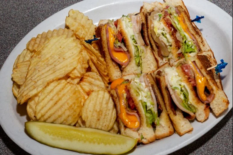 The club sandwich was first served in gentlemen’s clubs back in the late 1800s. The two-layer sandwich filled with chicken or turkey, bacon, lettuce and tomato layered between three slices of mayo-coated toast held together with a toothpick is elegant enough to be considered a knife-and-fork dish by some in the private club set. The version at Gadwall’s Grill is wholly satisfying and anything but stuffy. Three slices of bread are brushed with melted butter and tossed on the flattop. So is a slice of deli turkey, which is grilled with a slice of Swiss cheese. Same with deli ham and American cheese. Once both pieces of cheese start to melt and the bread is nicely toasted, it’s all stacked together with lettuce, pickle and mayo, cut into triangles and held together with frilly toothpicks.