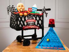 Competitors Sally Mach and Sherry Mach's cake large grocery cart with Guy's head inside and classic grocery items, in front of the cart is the pyramid game with a red light on top, during the Cake Challenge, as seen on Winner Cake All, Season 1.
