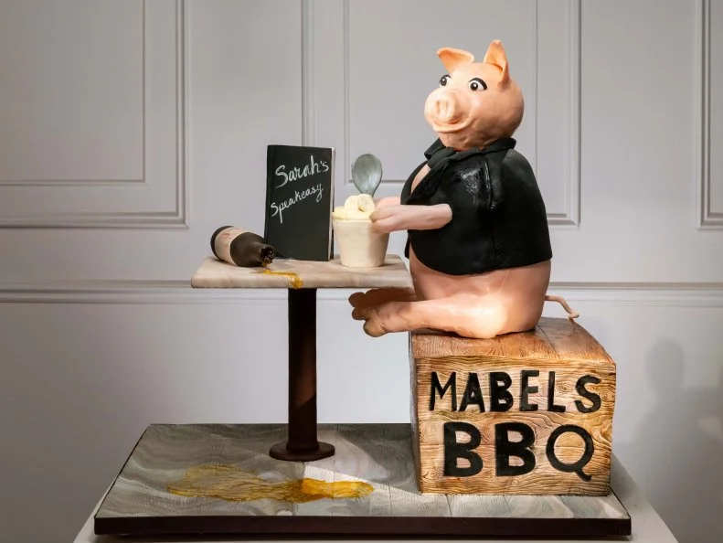 Competitors Rachael Louw and Samantha Petry's cake is A Lovely pig enjoying ale and banana pudding sitting on a crate from Maples BBQ, featuring a British denser cake banana, layer of sour cream chocolate ganache, mascarpone with caramelized pecans, banana foster top layer, mascarpone buttercream on the outside, during the cake challenge, as seen on Winner Cake All, Season 1.